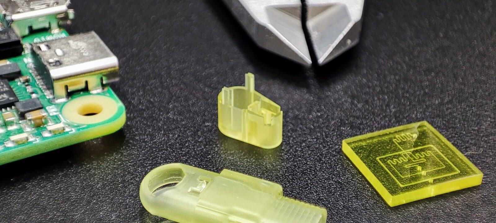 A variety of different small yellow parts created with micro 3D printing, with plyers and a circuit board for scale.